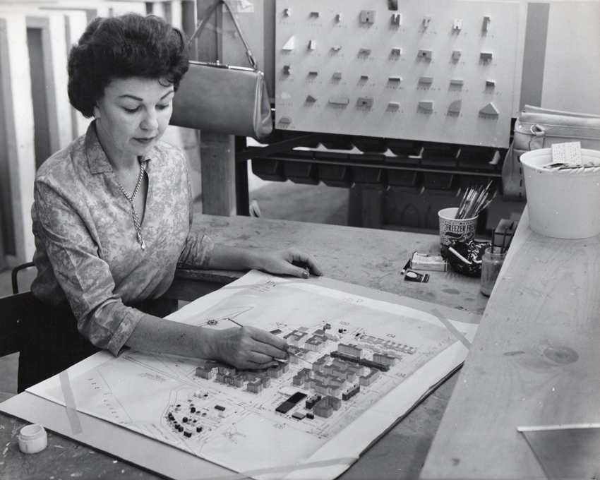 A woman from the 1960’s is carefully painting the model buildings on one of the panels that make up the large Panorama. On her desk are paintbrushes in a cup and miscellaneous tools. To her left is a board with various small shaped model pieces.
