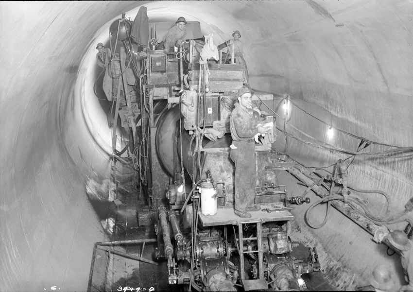 Four men stand on an industrial machine in a tunnel. They are dressed in boiler suits and hard hats.The machine is large and appears cumbersome.