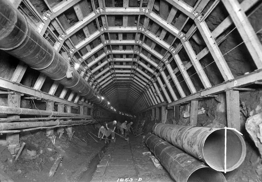 Inside the entrance of the aqueduct, large pipes lay to one side of the tunnel, one large pipe is on the wall,along with the surrounding metal infrastructure. Deep in the tunnel men with long ax picks and shovels working.