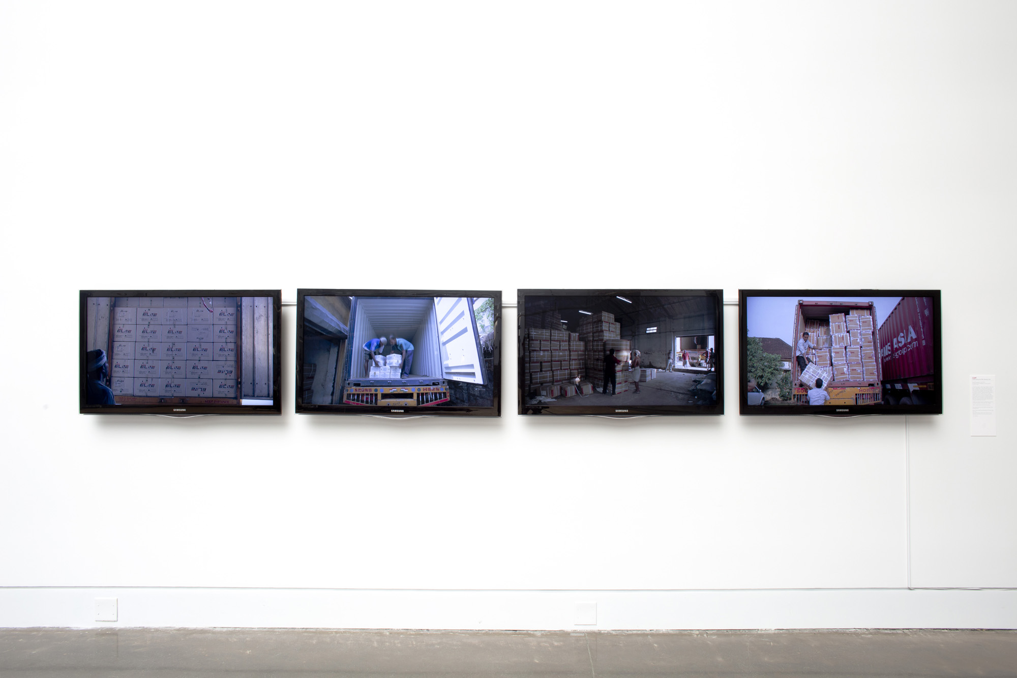 Four monitors installed in a row, on a white exhibition wall. The monitors are displaying scenes between a warehouse and a loading truck.