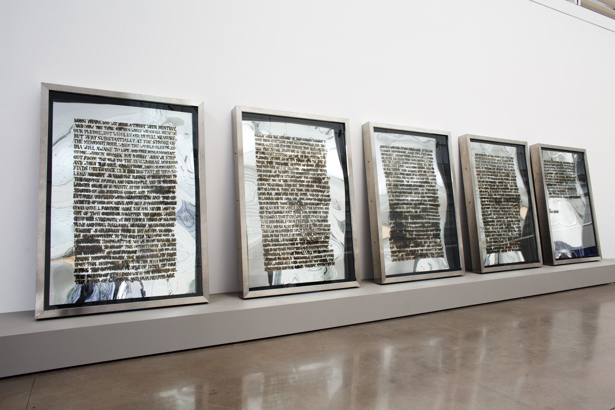 An exhibition wall with five-framed acrylic mirrors propped up against the wall on a skinny, gray platform. Each mirror has a block of text in black that’s illegible because of burn marks.