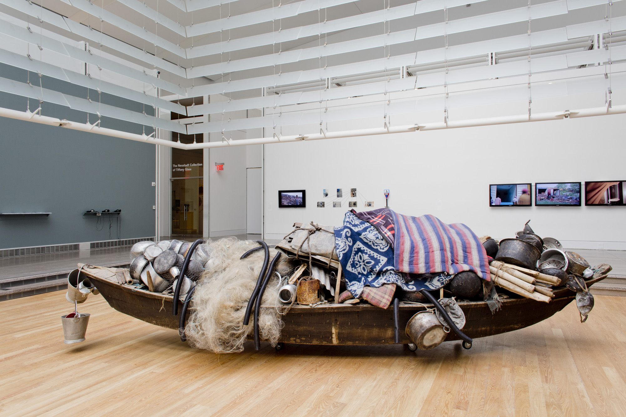 A found object sculpture installed in a hardwood exhibition floor. The sculpture is made of a weathered boat, overflowing with cooking pots, utensils, fabric, steel, a fishing net, bamboo, rope and a plastic pipe.