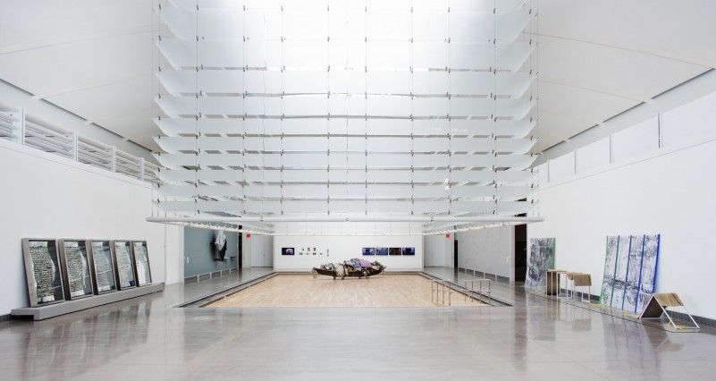 An exhibition space with artworks on the perimeter walls. At the center of the space is a set of four-sided, white blinds raised to reveal a section of hardwood floor embedded into the exhibition floor.