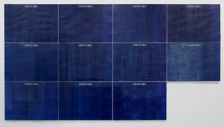 Installed on a white exhibition wall are eleven panels of paper saturated by blue, ballpoint pen. At the top of each piece of paper, in white serif font reads “ONONIMO”. Each panel has different line quality and texture.