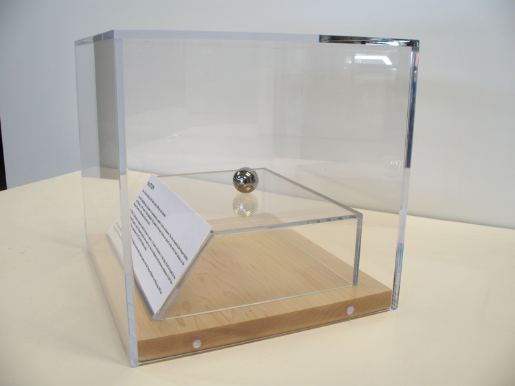 A clear, acrylic display case placed on an exhibition podium. Within the case is a small stand, also made of clear acrylic, with a stainless steel ball placed on it. On the side of the small stand is a white sheet of paper with text.
