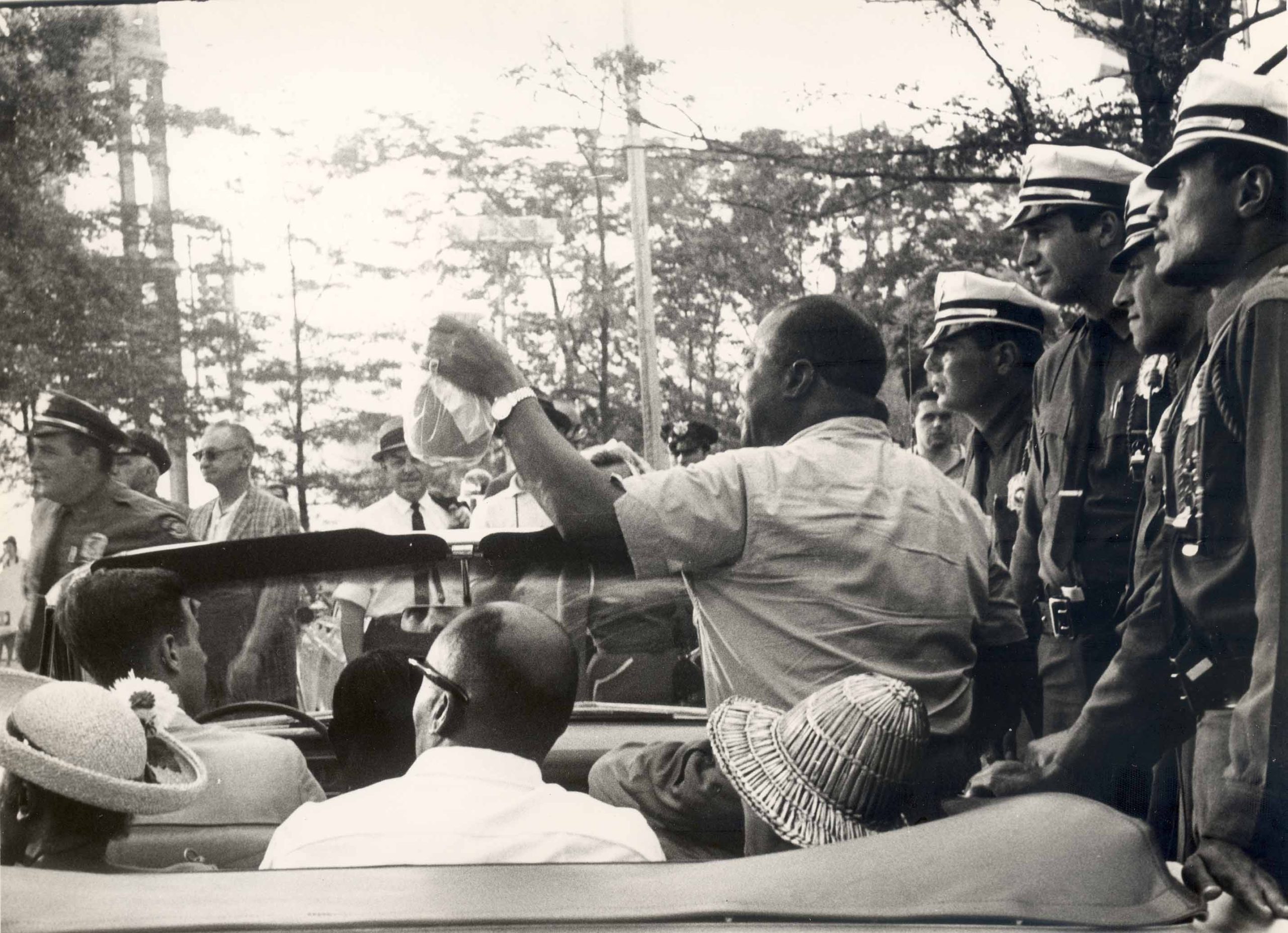 A black and white photograph of Louis Armstrong sitting up in a parked convertible filled with seated passengers. His back is turned away to wave to a crowd. Lined up against the convertible is a row of police officers.