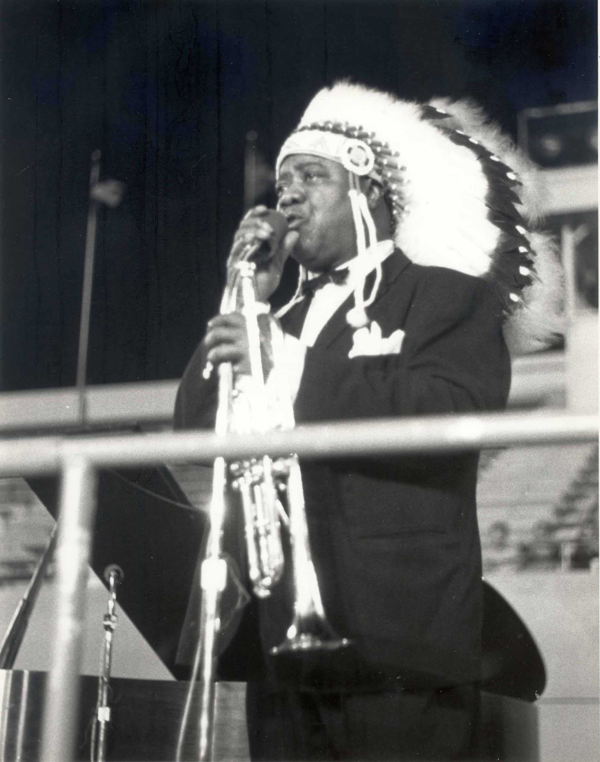 A black and white photograph of Louis Armstrong at a microphone. His hands are clasped around the microphone and stand, and he has an earnest expression on his face. He is dressed in a suit and wearing a Native American headdress.