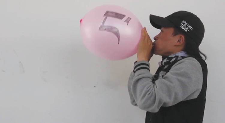 A video still of a man standing in front of a white wall. He is facing to the left and blowing up a pink balloon with his mouth. The balloon has a symbol sharpied on it. He is wearing a black and gray jacket, and a black cap with the phrase “why not? 1. 2. 3…2012” embroidered on in white.