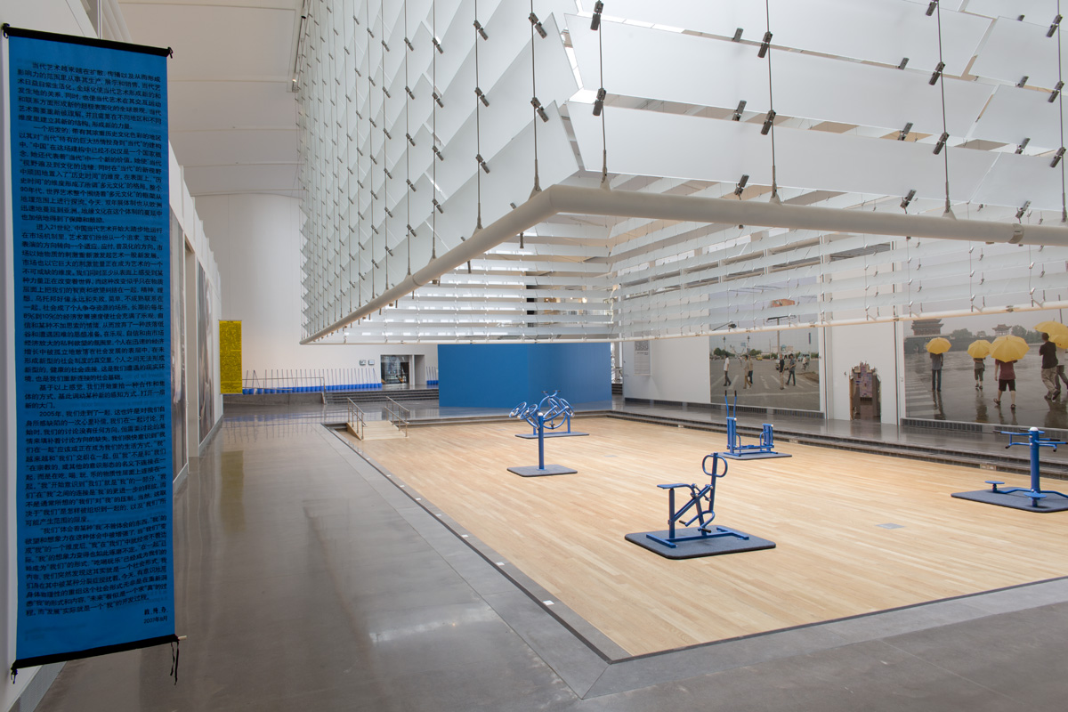 A white, four-sided set of blinds raised to reveal a hardwood floor, embedded into the concrete floor of an exhibition space. On the hardwood floors are five pieces of blue fitness equipment.