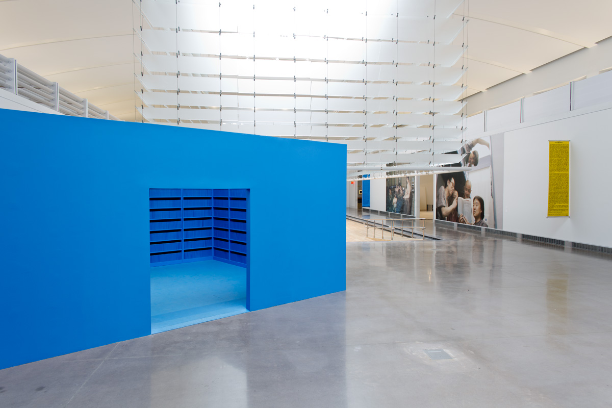 An entirely blue room installed on the floor of an exhibition space. The room has one entry way and inside the walls are made up of blue shelves filled with books.