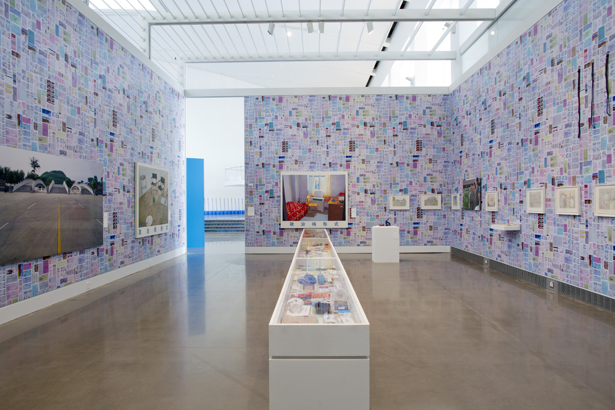 An exhibition room with walls covered in busy, purple wallpaper. Displayed on the walls are a series of large photographs and smaller artworks. In the center of the floor is a long and skinny display case.