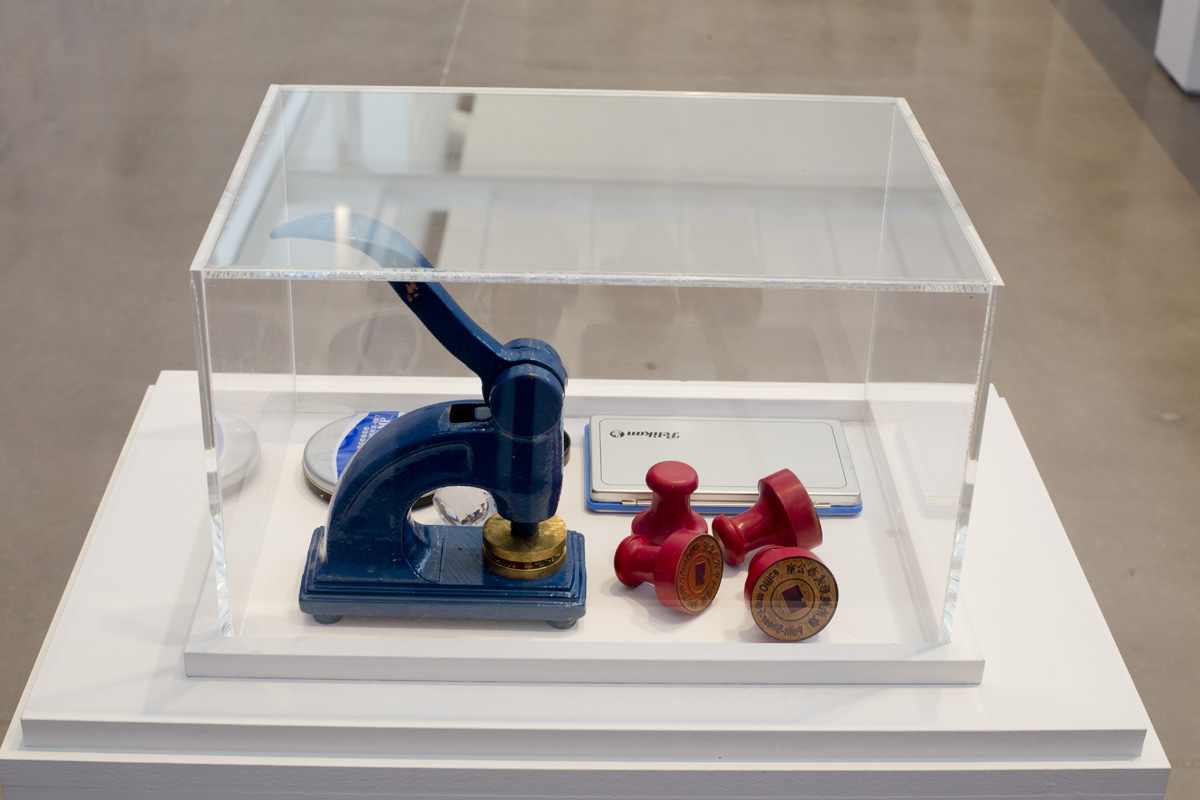A podium housing a glass display box. Inside the box are four red stamps, an ink pad, and a button maker.