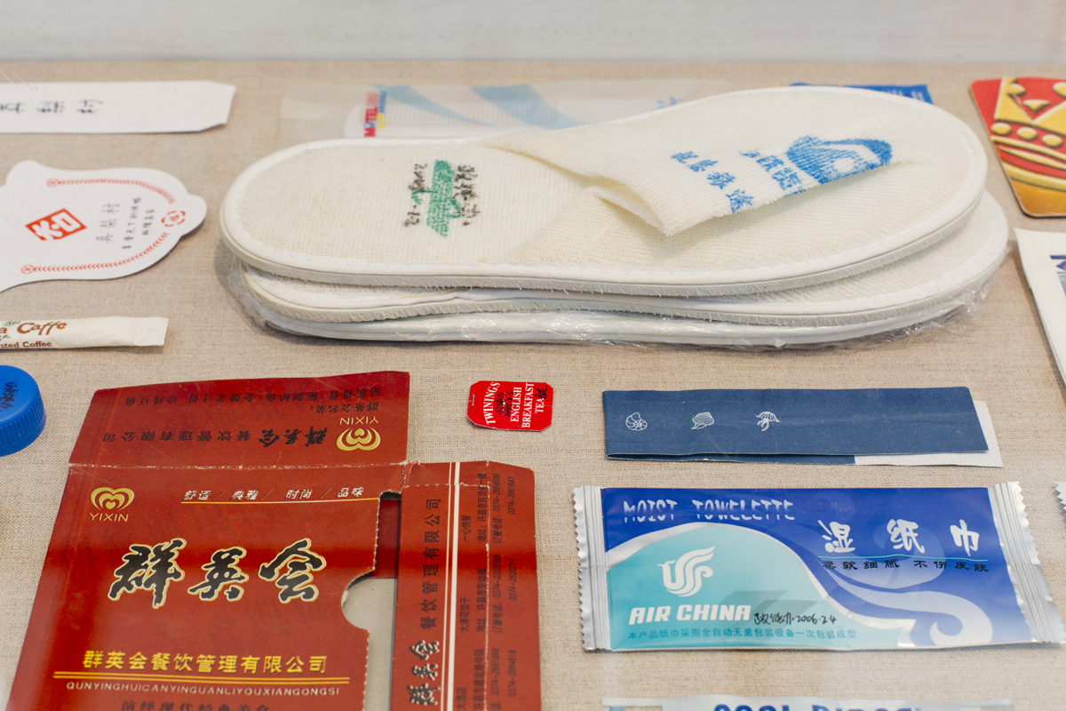 A close up of Chinese products and their wrappers laid out flat on a surface. Included in the collection are white slippers, an Air China moist towelette wrapper, a tea bag tag, a coaster, a plastic bottle, and a flattened out box.