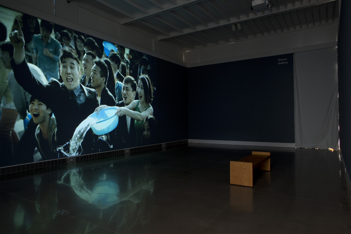 A dark exhibition room with a viewing bench placed on the floor. Covering an entire wall is a video still of a crowd of Chinese people cheering. One woman in the crowd is spilling water out of a baby blue, plastic tub.