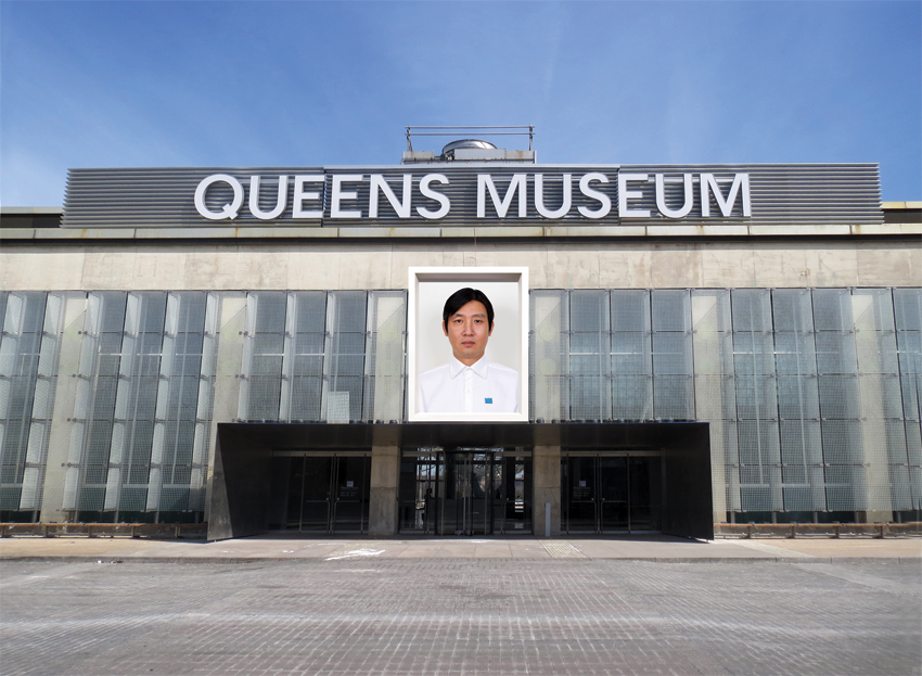The entrance to the Queens Museum against a blue, day-time sky. “Queens Museum” is spelled out in silver signage across the top of the building. Hanging below the sign and above the front doors is a color photograph portrait of a Chinese man. He is posing against a white backdrop and wearing a white button up.