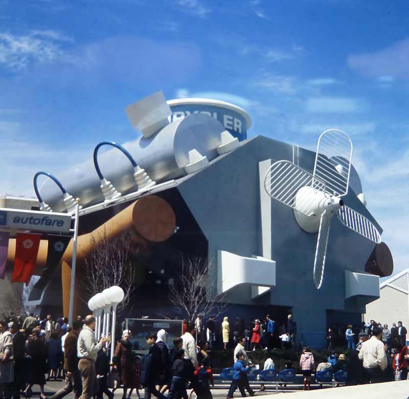 A color photograph of a pavilion shaped like an engine. Notably there is a giant, white, propeller on the front side of the building. Down at the street is a crowd of visitors.