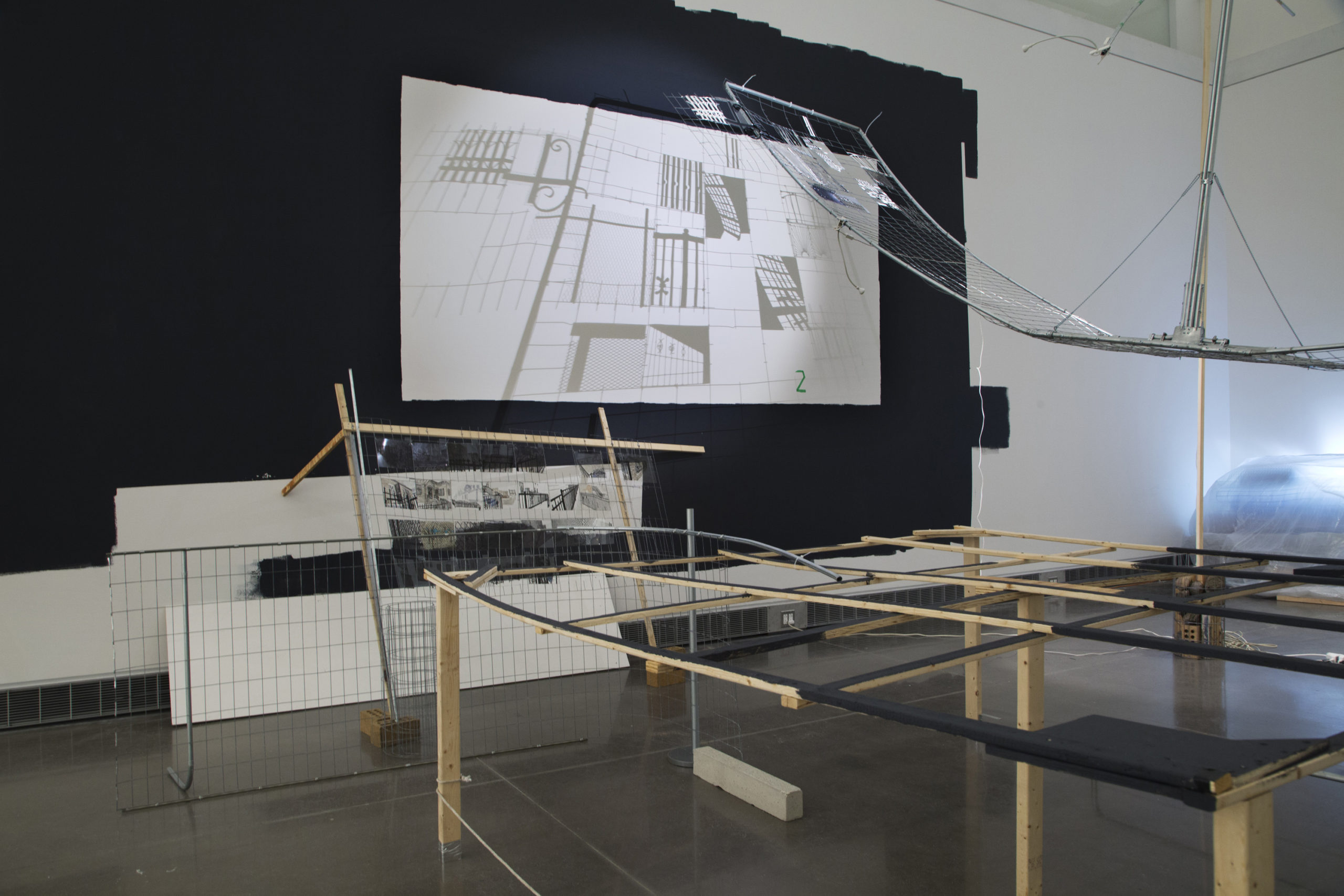 A sculpture installation made up of construction materials including thin pieces of wood, black fabric, metal pipes, and fencing. The materials are loosely linked together to create uncontained spaces. In the background the exhibition wall is covered with black fabric. Layered on the fabric is paper with a simple gridded drawing similar to the patterns found in the sculpture installation.