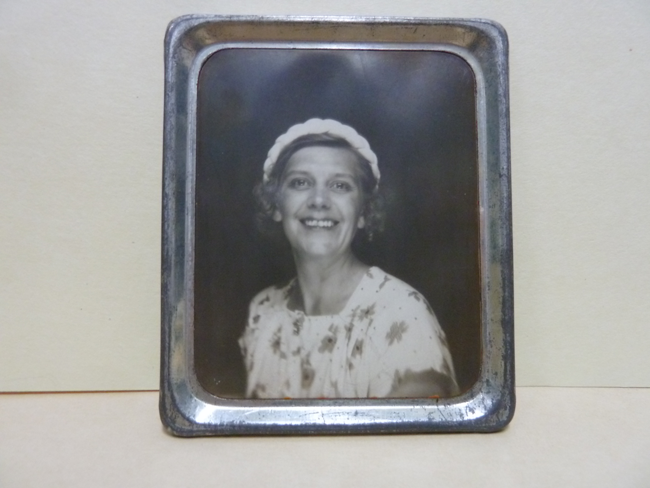 A black and white portrait of a woman in a metal frame. The woman has fair skin, short curly hair and a kind smile. She is wearing a floral top and a headband.