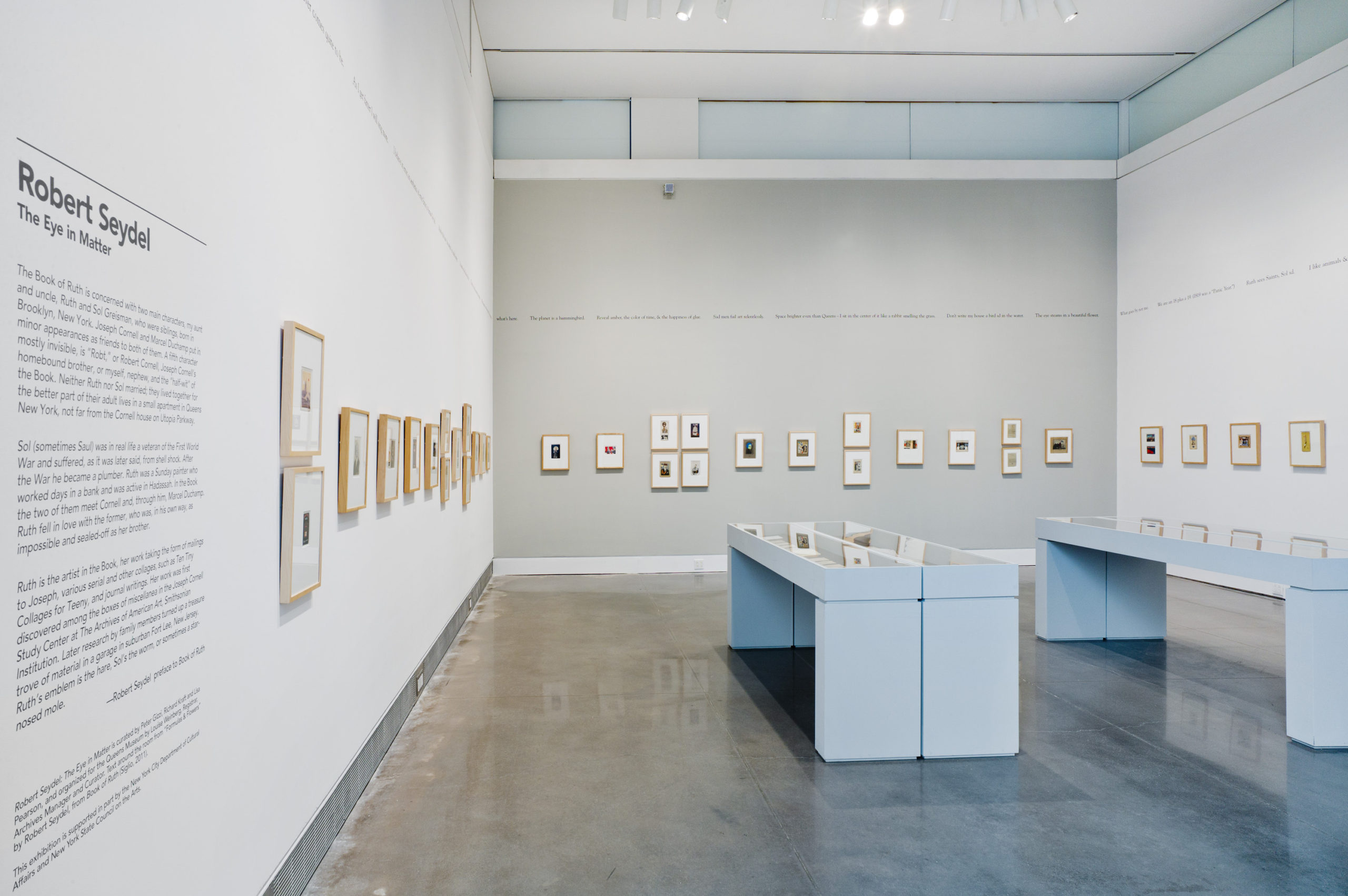 An exhibition room with white and gray walls. Starting on the left wall is a block of exhibition text, the featured artist’s name, followed by the show title; “Robert Seydel, The Eye in Matter”. Following the wall text is a series of framed and matted collages that wrap around three walls. On the exhibition floor are two white display cases.