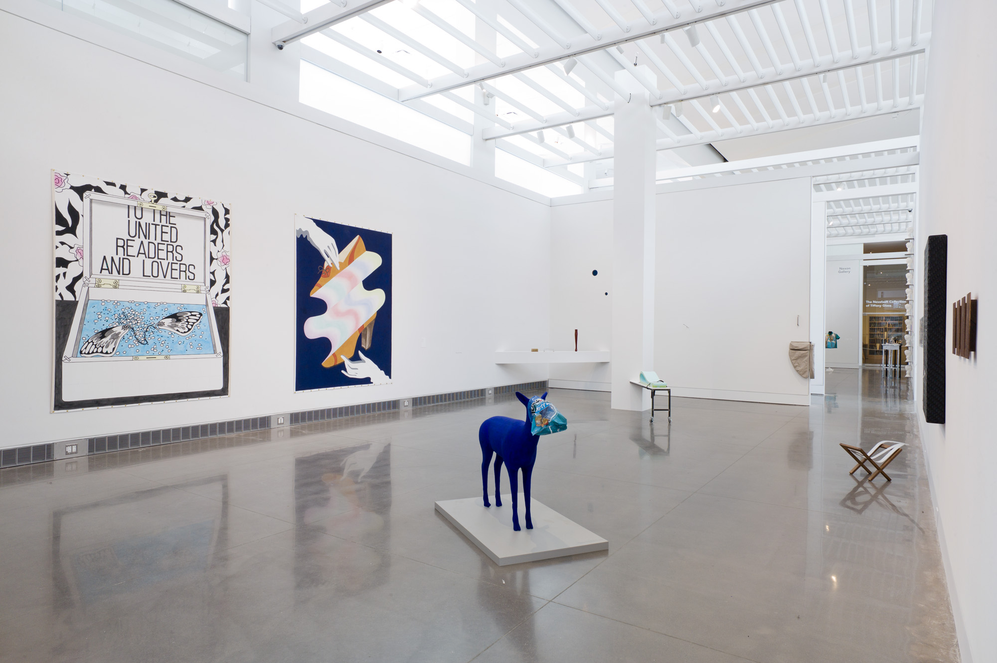 An exhibition room with large, two-dimensional artworks and sculptures hanging from the walls. On the exhibition floor is a sculpture of a dark blue, four-legged animal. The animal’s face is covered by a blue Tostitos chip bag.