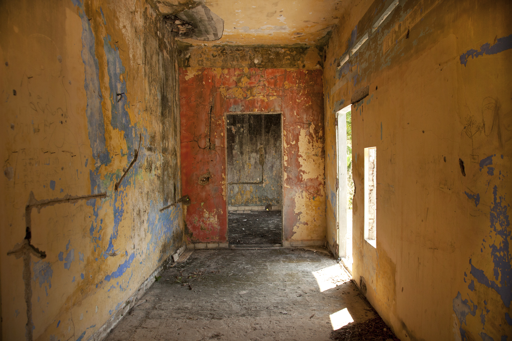 A photograph of an interior space with peeling, cracked wall paint and de-tiled floors. The wall paint is a warm yellow, with swashes of red and blue. On the furthest wall is an entryway leading to another room. On the left wall is another entryway that leads outside and a window. Sunlight is casting into the interior from both the window and entryway.