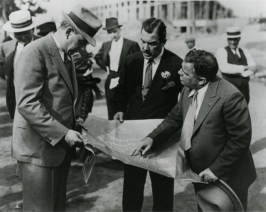 A black and white photo of Robert Moses, Grover Whalen, and Mayor Fiorello La Guardia speaking over a map of the 1939-40s World's Fair.