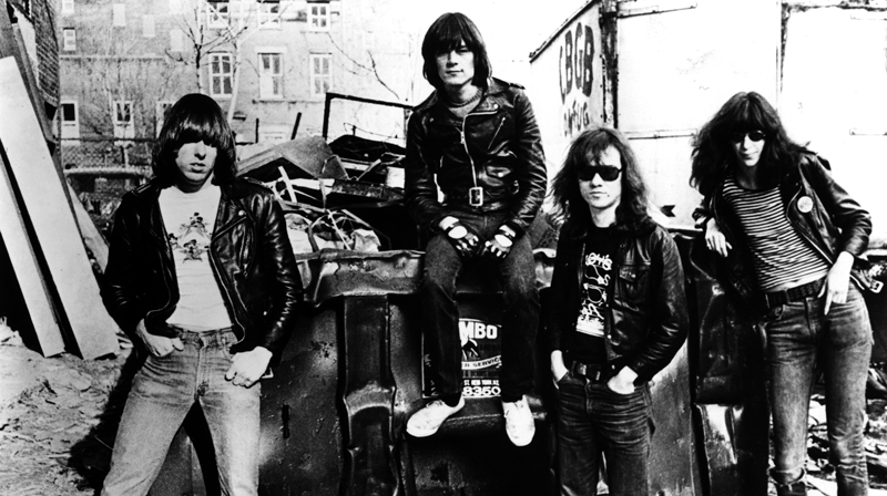 A black and white photograph of the members of punk band, The Ramones. The four band members are posing in an alley filled with junk. They each have long, shaggy hair with bangs, are wearing skinny jeans, t-shirts, and black, leather jackets. Each has a cool and tough expression on their face.