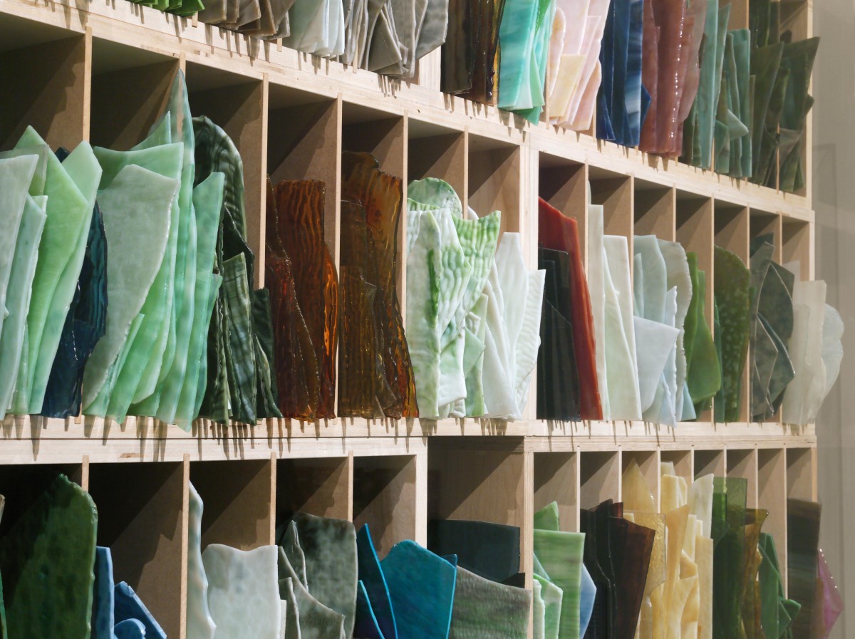 A blonde-wood shelf with cubby spaces filled with sheets of glass organized by color.