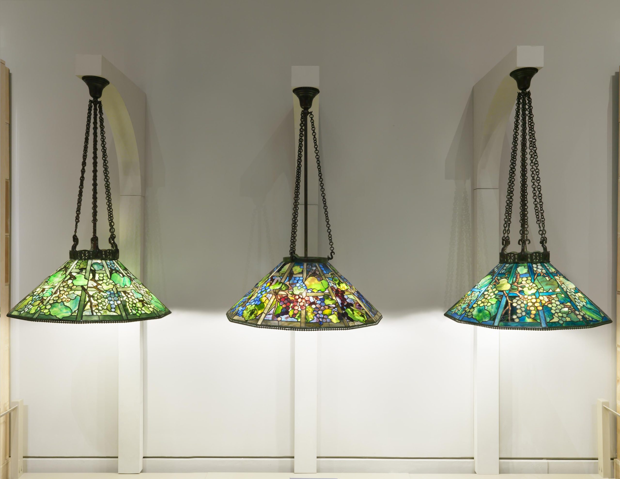 Three hanging Tiffany Glass lamps installed on an exhibition wall. Each lamp shade is hanging from a black, three-piece metal chain. The lamps are made of green, blue, and purple tones and depict grapes growing from a vine.
