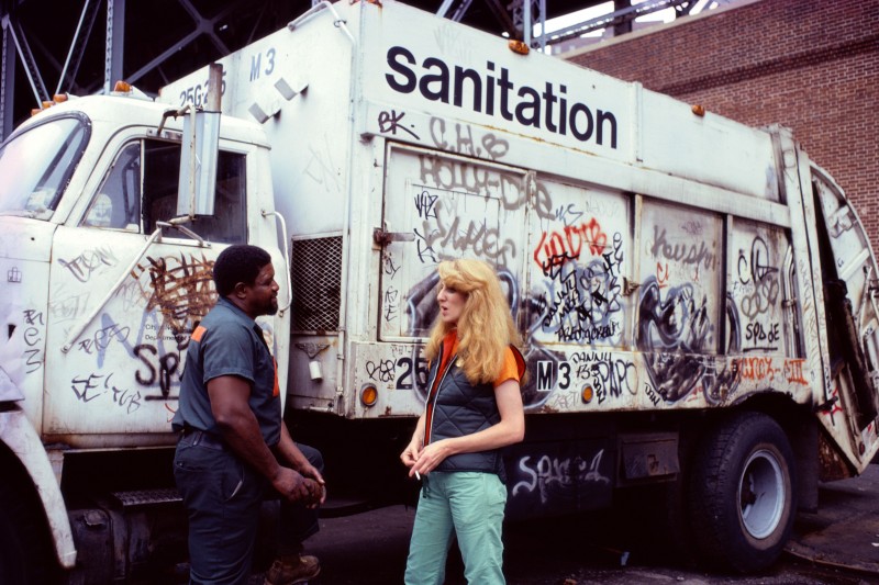 The artist Mierle Laderman in conversation with a Sanitation worker, standing beside a Sanitation truck. Mierle is a White woman with thick, long, blonde hair. She is wearing teal pants, an orange shirt, and a black, zip-up vest. The Sanitation worker is Black and dressed in a gray uniform. The truck is white and covered in graffiti.