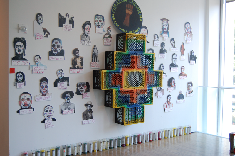 A white exhibition wall with milk crates hanging in a cross shape. The outer facing surface of the crates have been spray painted, starting from the center and radiating out, yellow, orange, white, blue, green, and yellow again. Around the cross are paper cut out images of community members and famous people wearing La Calavera face paint. Propped up on the milk crate cross is a black, round sign with a brown power fist and the phrase “Corazón Indígena Por Las Vida Negras”. Below, at the gallery floor, is a row of chakra candles in different colors.