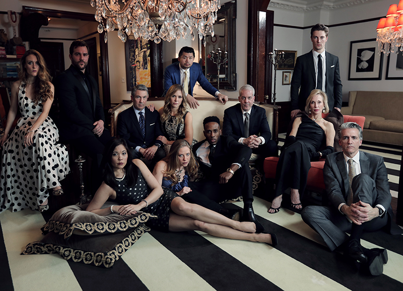 Twelve adults of different genders and racial backgrounds pose for a group portrait in a swanky living room scene. The adults are dressed in suits and formal gowns. The room has black and white striped floors, a number of lounge chairs and sofas, framed art, diamond chandeliers and crown molding.