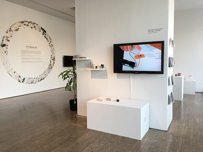 An exhibition space with a collaged, ring-shaped decal on the far left wall. Within the ring is the show title which reads “It’s About Us” and a block of wall text underneath. In the center of the floor is a white, center column housing a monitor, shelves, pamphlets, and small objects. Behind the center column is a set of podiums displaying burnt sienna and white colored sculptures.
