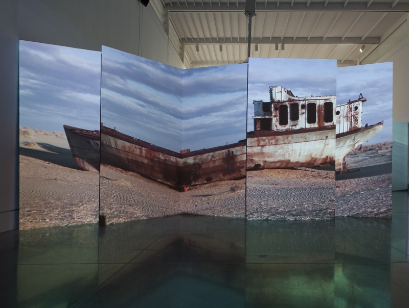An image of a rusted ship sitting on a sandy beach, close to the water and against a cloudy sky. The image is split between four large-scale panels installed in the middle of an exhibition floor.