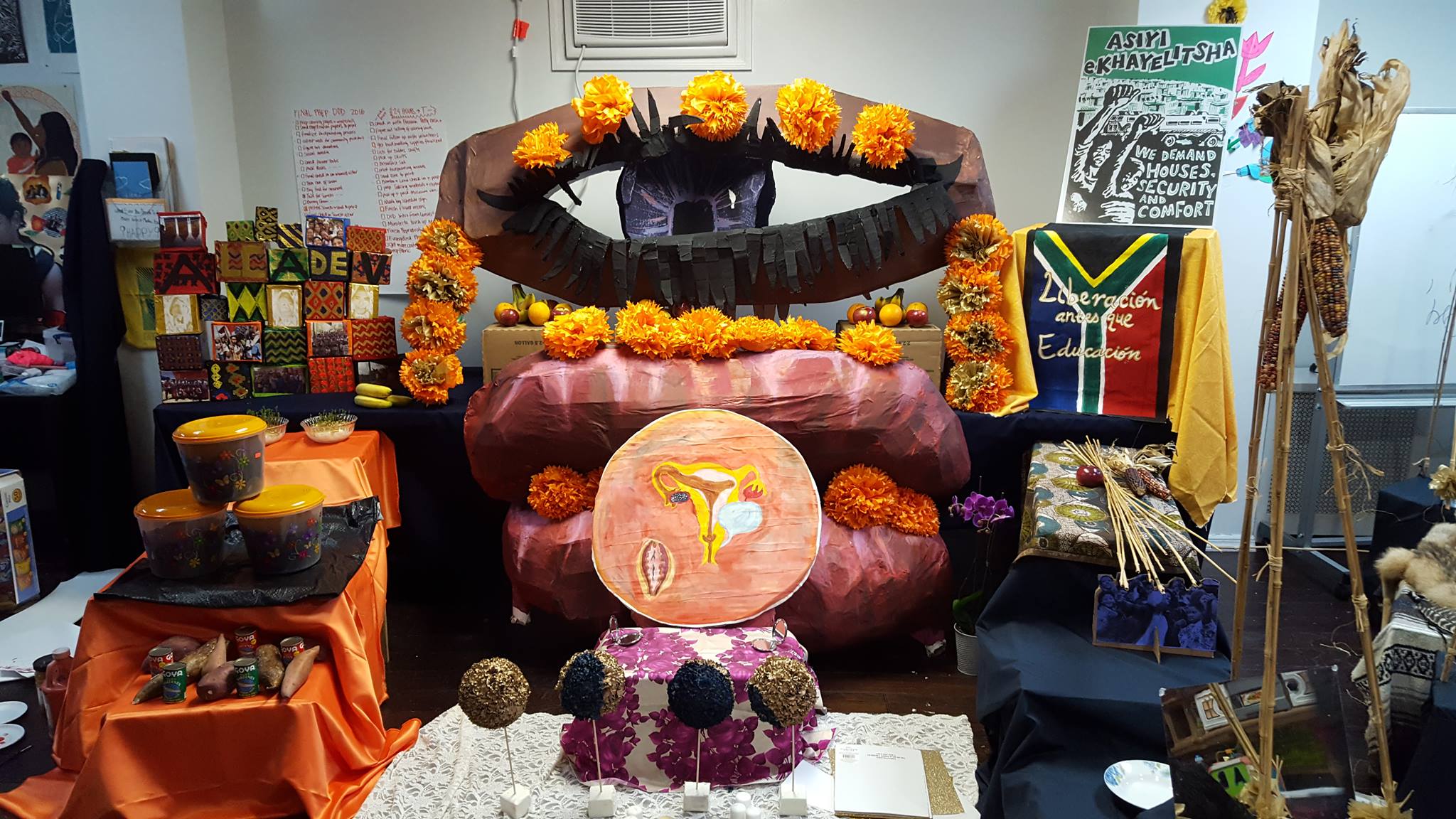 An altar space with a large papier-mâché eyeball, pair of lips, and circle with an illustration of a uterus. The papier-mâché sculptures are adorned with orange flowers. On each side of the sculpture are staircased stands, covered with orange and navy blue fabric. On each stand are food offerings like corn, Goya cans, tupperware, and fruit. Also included is a flag that reads “liberación antes que educación