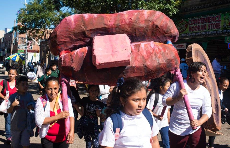 A group of Latinx women and children march through a neighborhood street, carrying large papier-mâché lips above their heads.