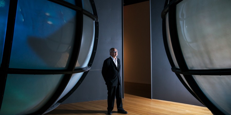 An elderly Chinese-American man stands in a room with hardwood floors and dark grey walls. The view of him is framed by two large, iridescent globes that are covered by black, gridded lines. He is wearing a black suit, that fits his body loosely, a white dress shirt, and glasses.