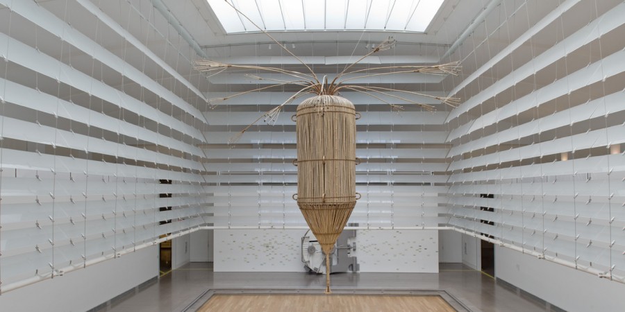 A large-scale, funnel-shaped sculpture fabricated from a natural fiber. The sculpture is hanging from the ceiling, within a four-sided set of blinds that have been raised to reveal the exhibition space below.