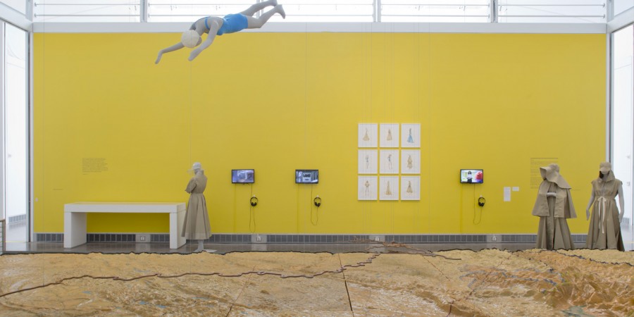 A relief map of New York City sits in front of an exhibition wall. The exhibition wall is painted yellow and has a series of works including a display case, three mannequins dressed in khaki colored jackets, three monitors with headphones, and three by three fashion design sketches on white paper, framed. Hanging from the ceiling is another mannequin, in a swim diving position, dressed in an old school one-piece bathing suit and cap.