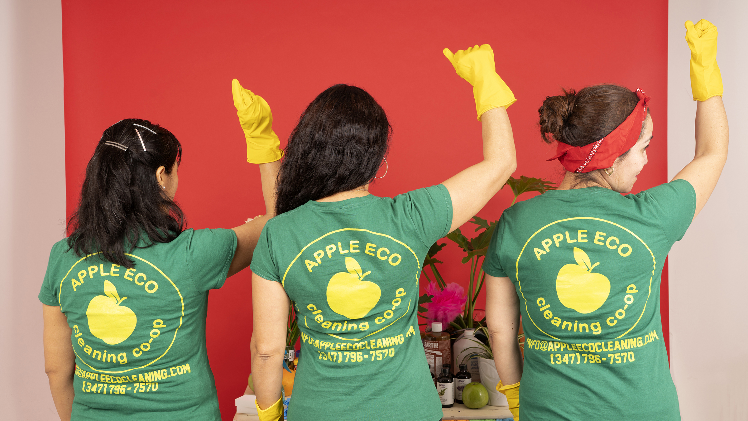 Three immigrant women stand in front of a red backdrop with their faces turned away. Each of them are wearing yellow cleaning gloves and are holding a power fist above their heads. On the back of their matching green t-shirts is a yellow apple with the words “Apple Eco cleaning co-op”circling the apple and sitting within a thin yellow circle. Below the circle, in the same yellow is the Apple Eco cleaning co-op’s email and phone number.