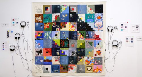 A quilt made up of 64 smaller square patches installed on an exhibition wall. Headphones with wires coming out from beneath the quilt, are pinned on each side. Along with exhibition text.