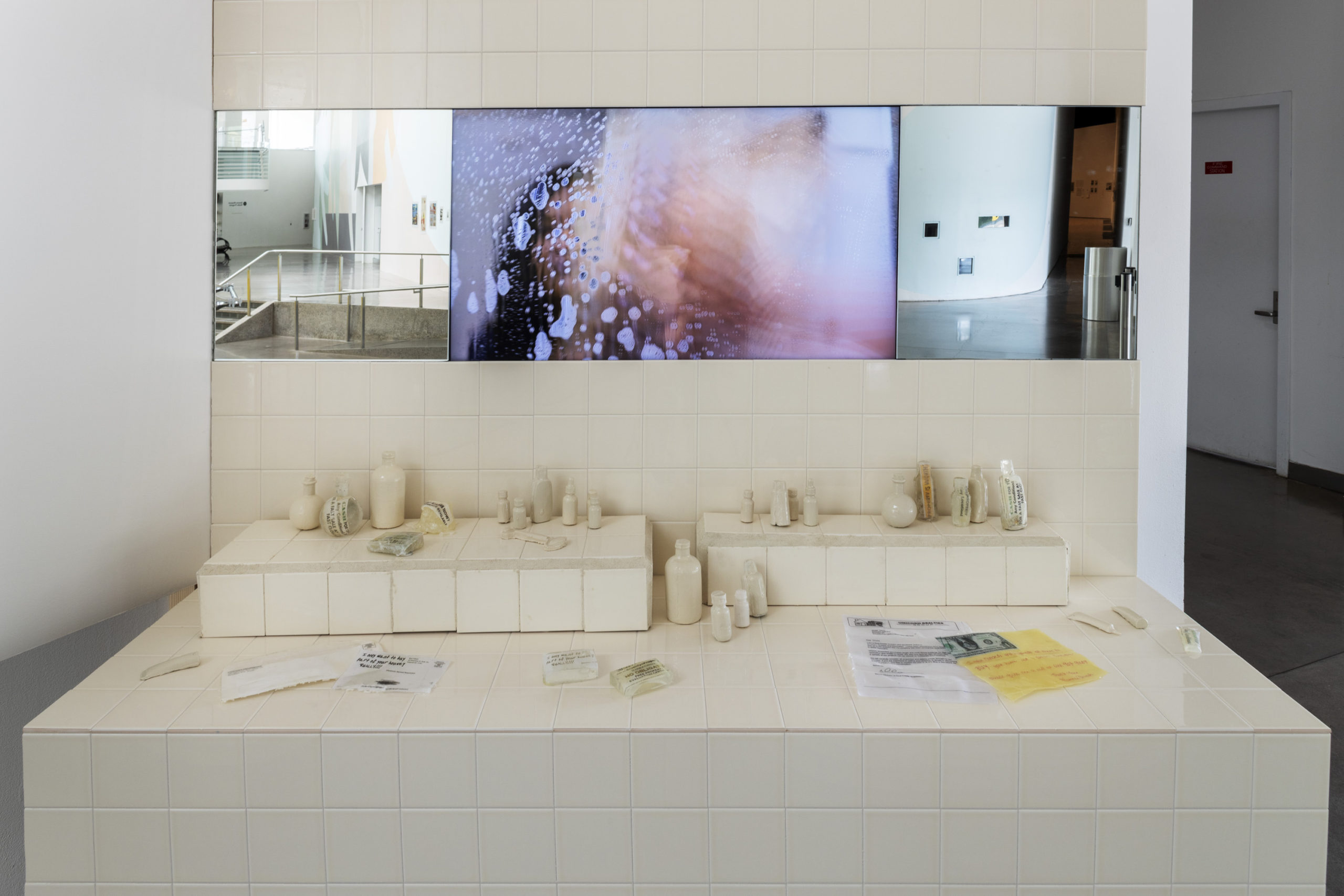 A cream colored tiled wall and counter surface installed on a white wall. On the wall is three images. The first and third are two images of bare exhibition spaces. The centered image is of a woman with a watercolor like wash distorting her likeness. On the counter are two stands made of the same cream colored tile, ceramic bathroom products, and a few pieces of paper.