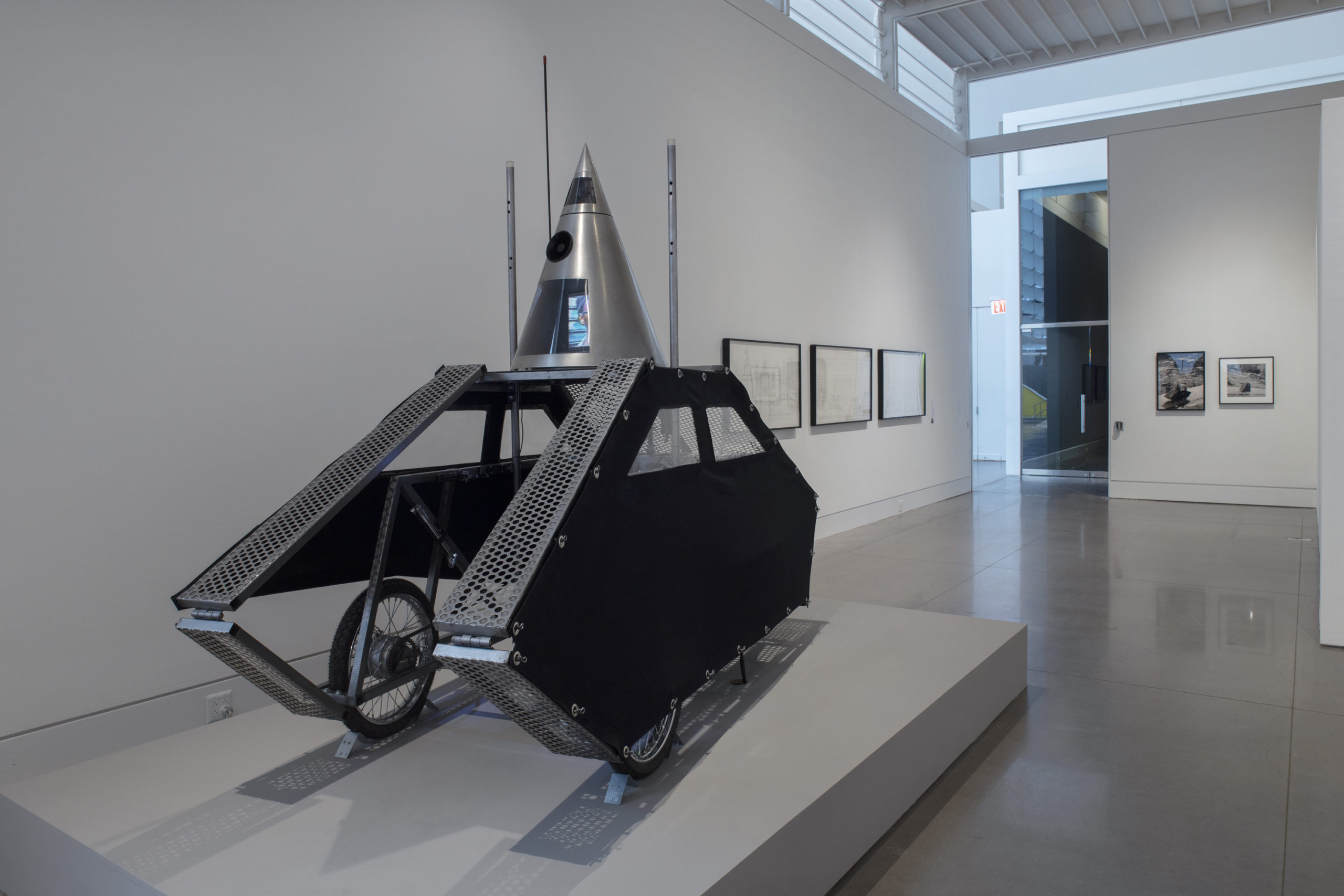 A white exhibition space with a large, low and wide pedestal installed on the floor. On the pedestal sits a sliver and black, mobile sculpture. It has two walls with wheels on the inside, and a silver cone at the top. In the back of the room are two rows of two-dimensional works on display.