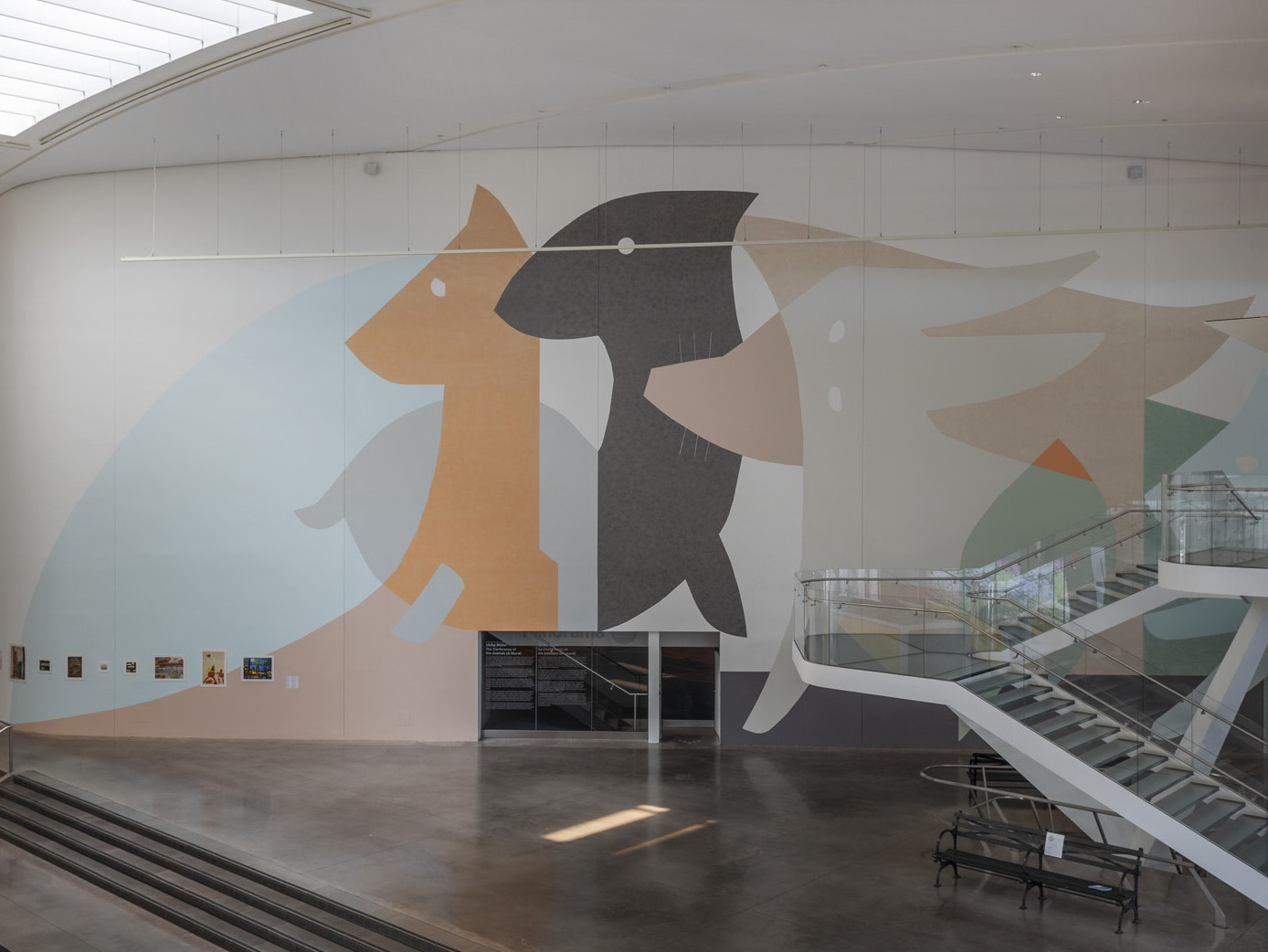A two story mural of abstracted animals in pastel and earth tones. At the bottom left of the mural is row of artwork on display. Descending from the top right is a twisting flight of stairs.