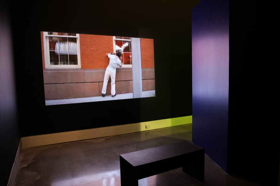 A black room with a black visitor bench placed in front of a large screen. Displayed on the screen is a still of a person, dressed in an all white outfit, peering into the window of an orange, brick building.