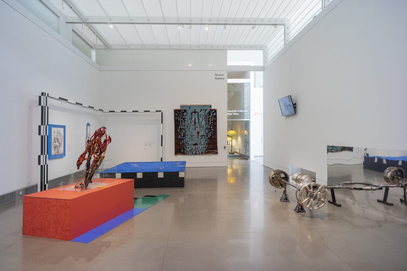 A wide installation view of Asif Main’s RAF: Prosthetic Location exhibition. To the right are three abstract compositions hanging on the walls. In front of the hanging pieces, are two large blocks, on the gallery floor. One is orange and one is black and blue. The orange block has an abstract orange sculpture on top of it. Framing the large blocks is a rectangular shaped beam with checkered black and white stripes. On the right hangs a flat screen, wall monitor and a propped up car axle on the floor.