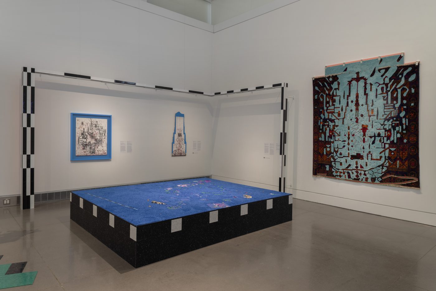 A corner installation view of Asif Mian’s RAF: Prosthetic Location exhibition. A large black block with a fuzzy blue surface sits on the gallery floor. Against the wall, on the left, is a rectangular shaped beam with black and white checkered stripes. Split between both walls are three abstract compositions of different sizes and museum text.