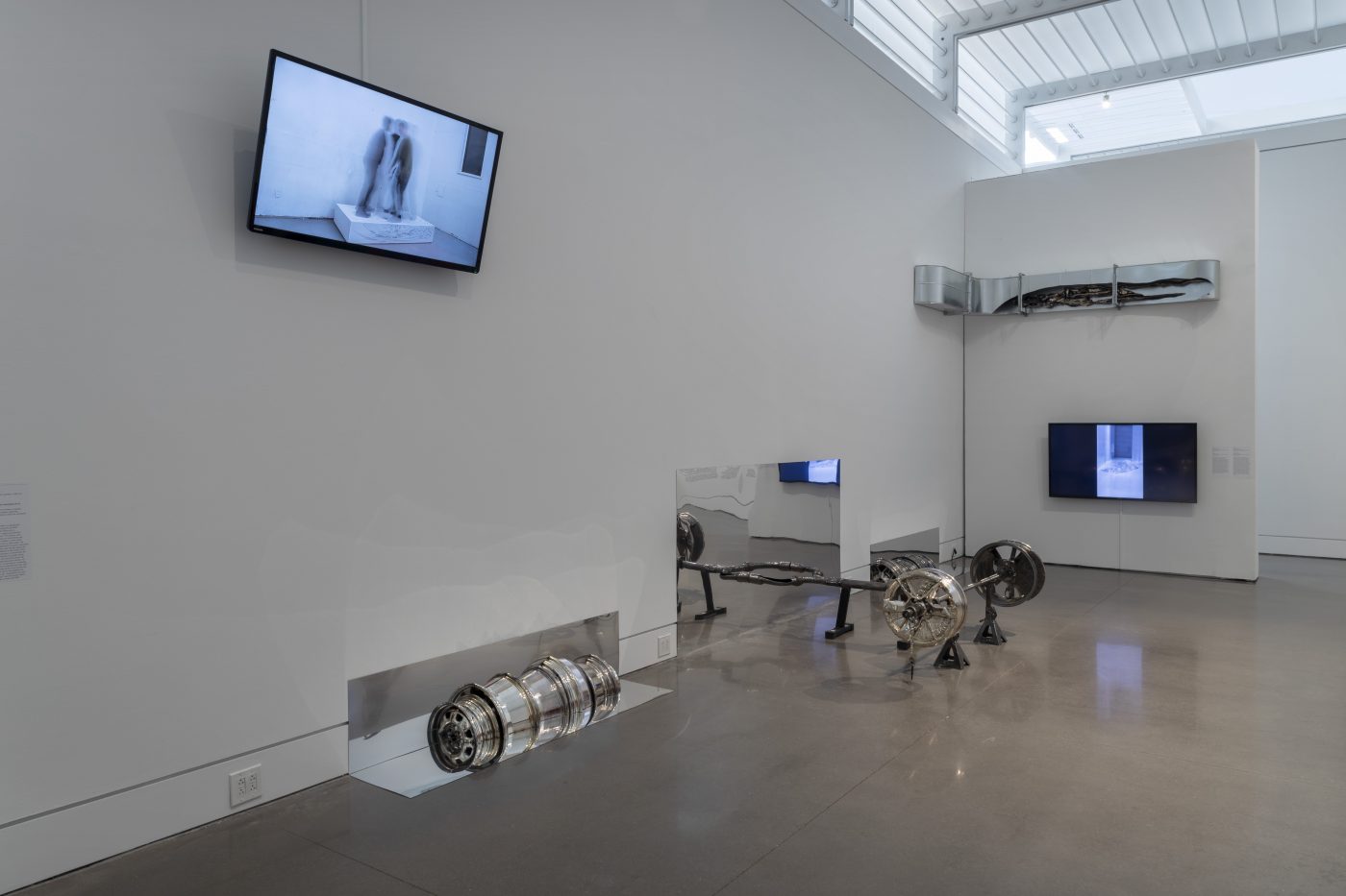 A corner installation view of Asif Main’s RAF: Prosthetic Location exhibition. On both walls hang flat screen monitors. In the corner is a thick chunk of metal, installed like a hinge. On the floor, leaned up against the longer wall are a trio of frameless mirrors. Placed in front of the mirrors are a row of rims and a propped up car axle.