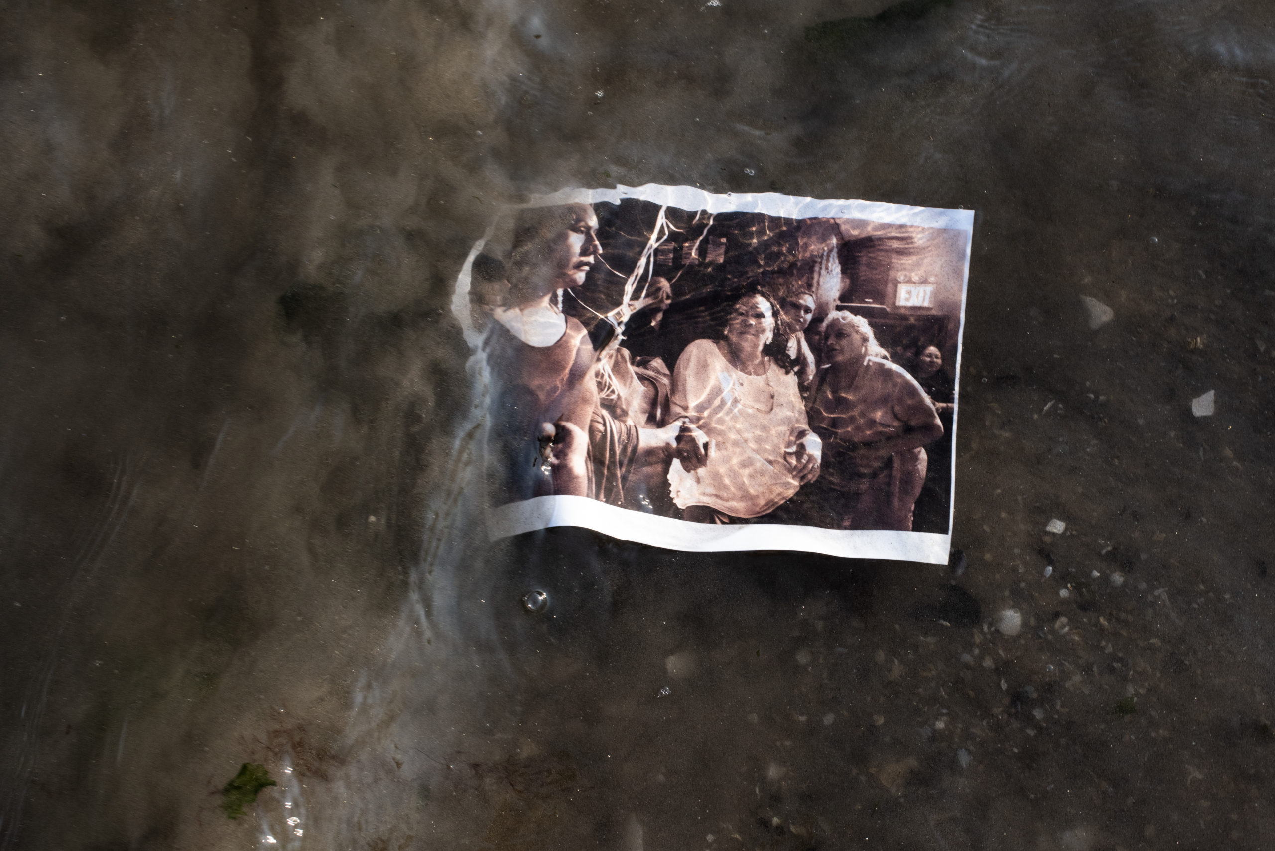 A polaroid photo, of a group of women of color, submerged in water. Ripples of water distort the clarity of the image. Beneath the surface of the water is brown asphalt and dirt.