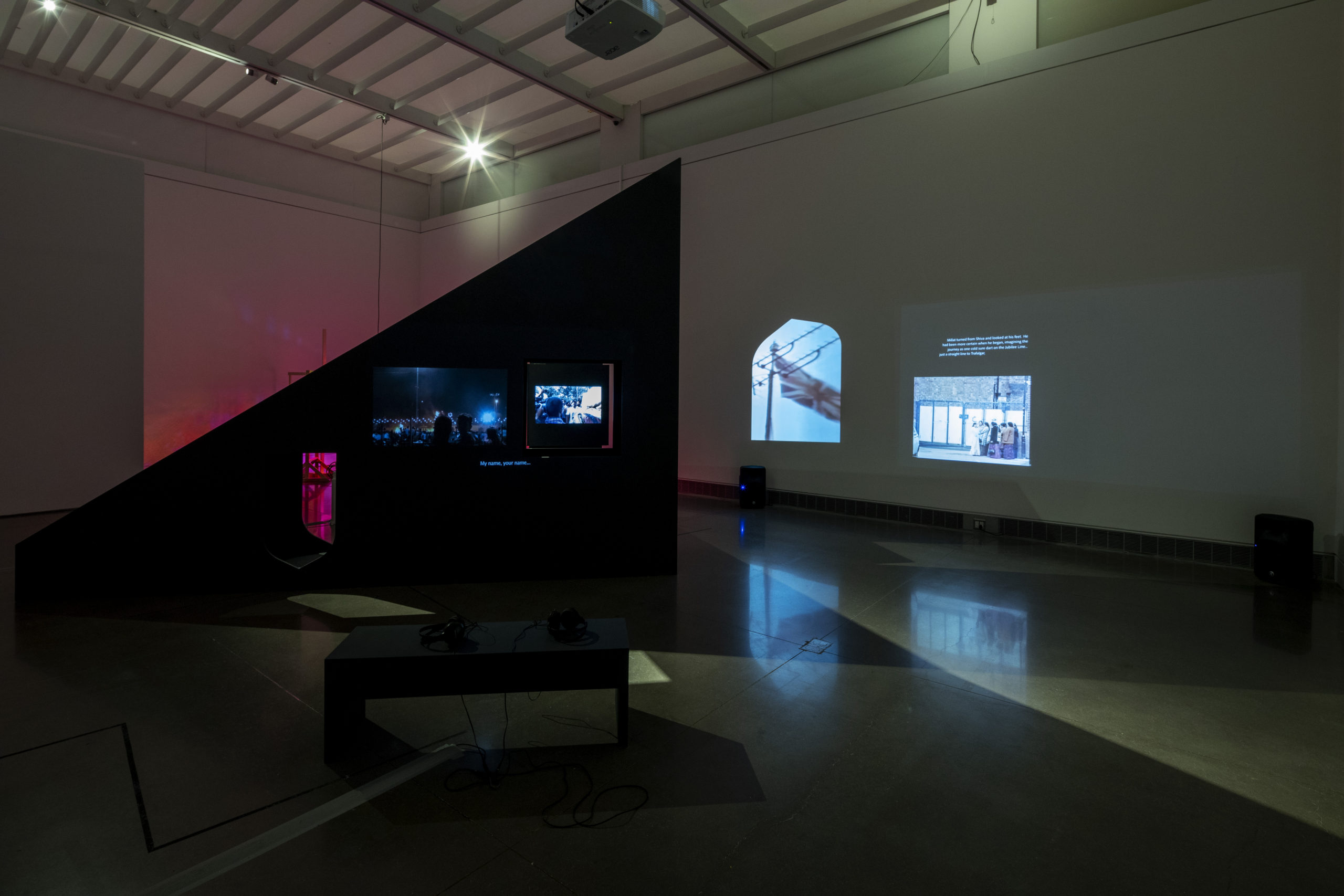 Behind a black bench with headphones is a huge triangular sundial structure containing two square cutouts through which protest video stills are projected. On the white wall to the right are two more projections: an archival Union Jack flag, and small white text above a still of a group of South Asian women on a labor strike.
